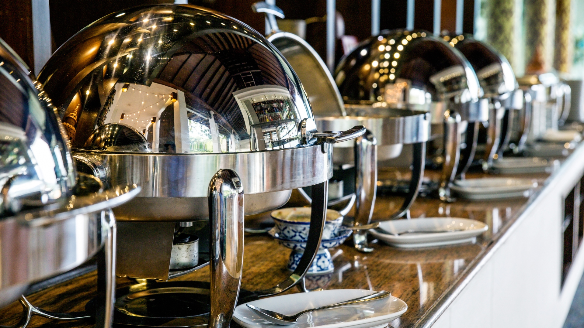 Buffet with shiny metal chafing dishes and plates aligned on a countertop.