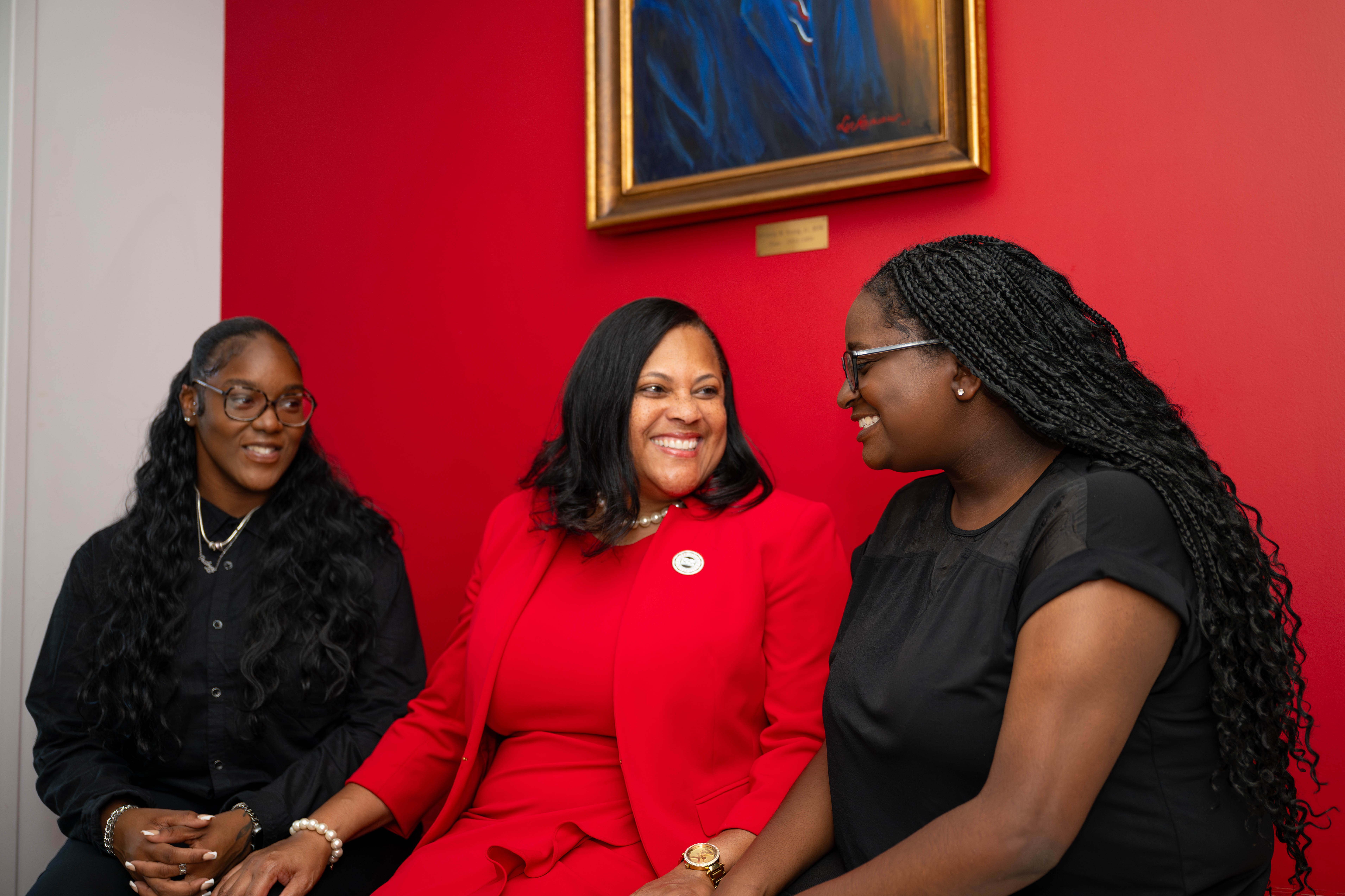 Three women seated, smiling, against a red wall with a framed painting.