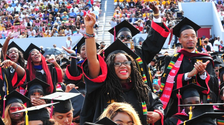 Graduates in caps and gowns celebrate, raising hands joyfully in a crowded stadium.