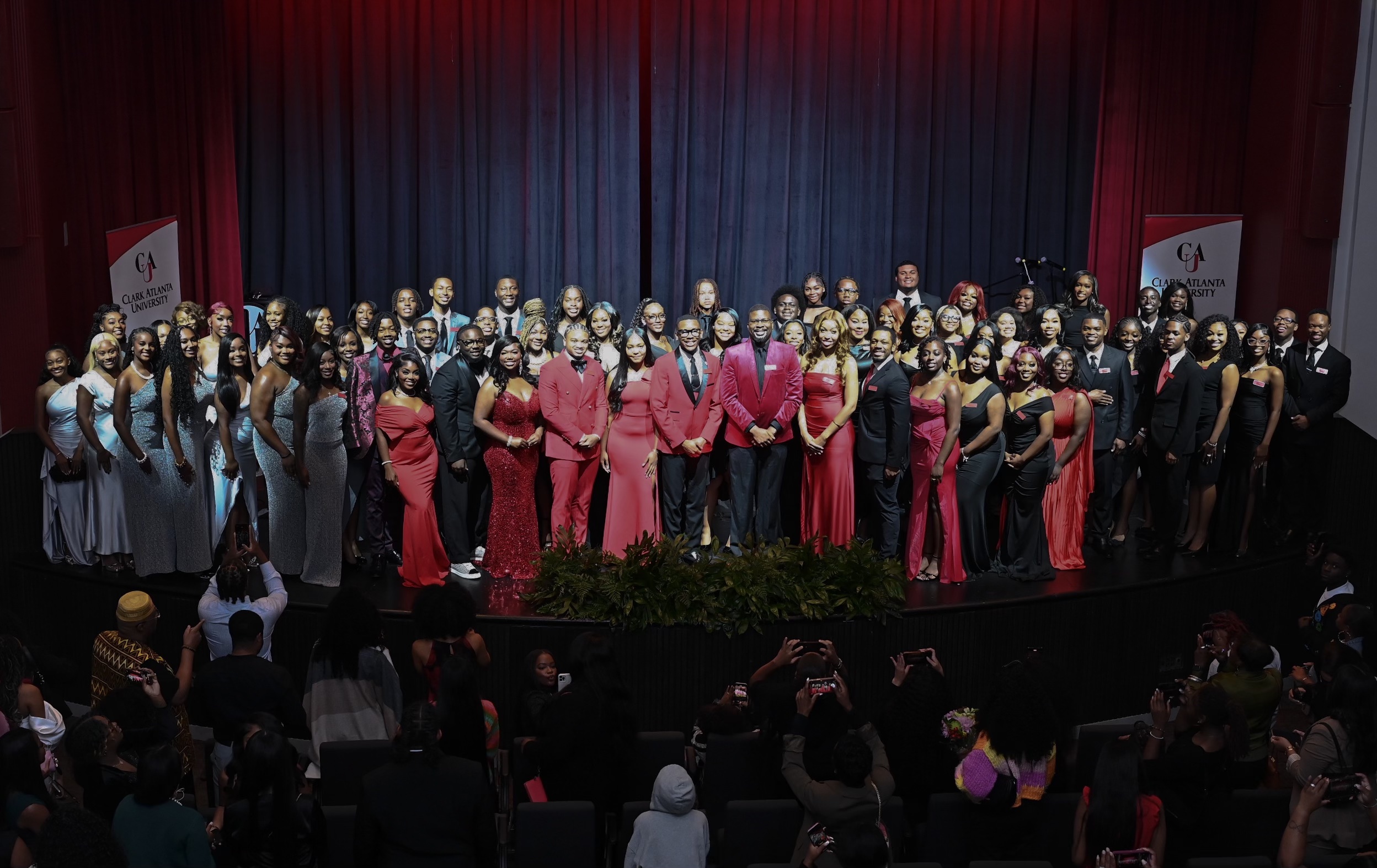 Large group in formal attire on stage, wearing suits and gowns, against a dark curtain backdrop.