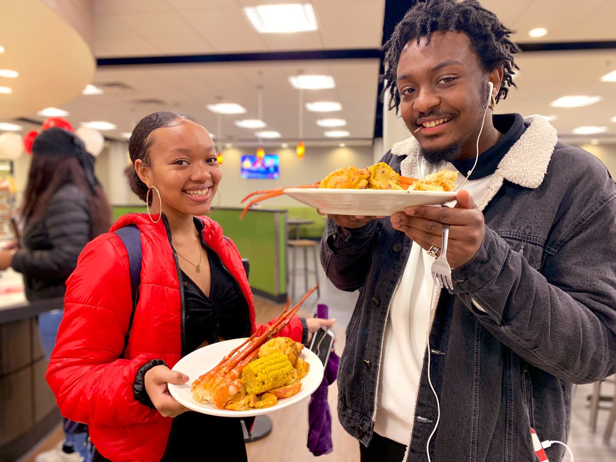 Two people smiling, holding plates of food in a restaurant setting.