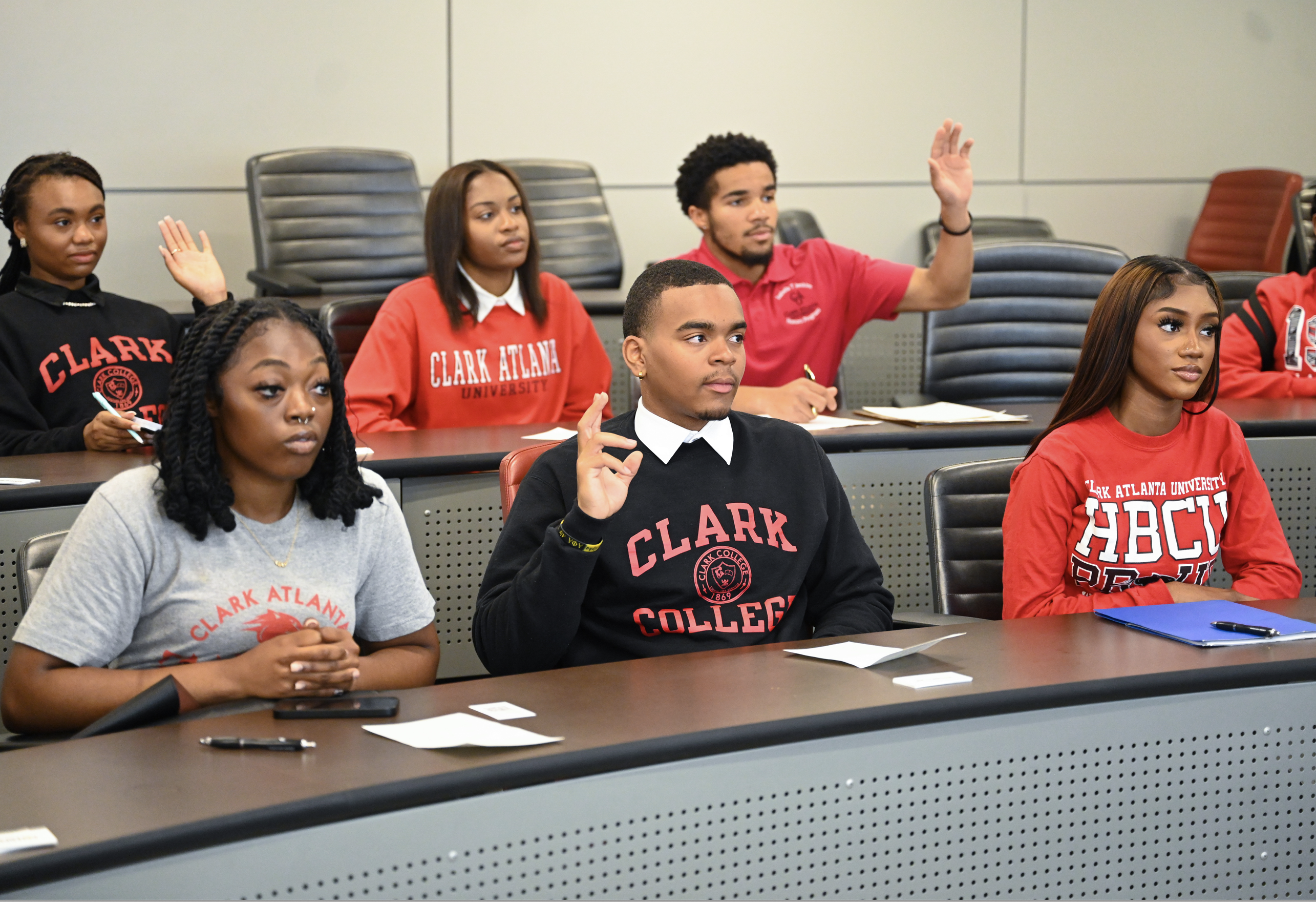 Students in a classroom, some raising hands, wearing collegiate sweatshirts.