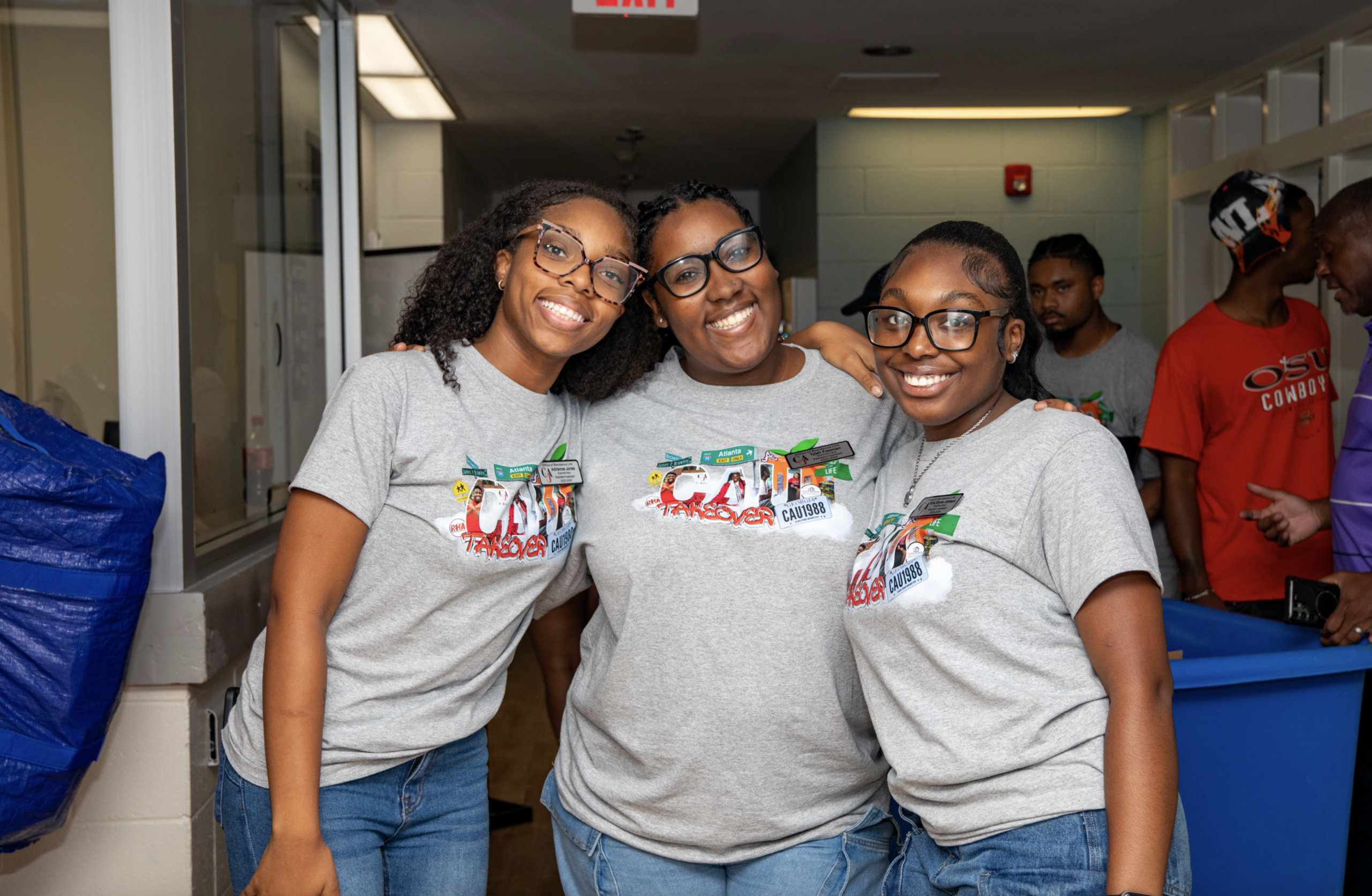Three smiling women in matching gray t-shirts stand together indoors.