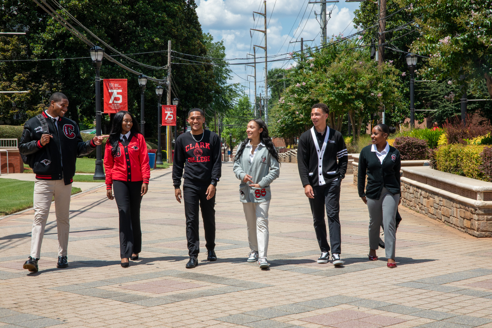 Six people walking on a sunny campus path, trees and banners in the background.
