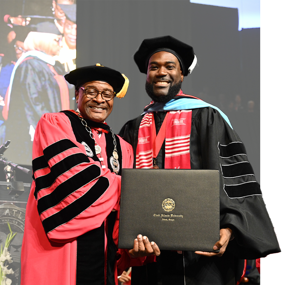 Graduate and faculty member in gowns holding a diploma on stage, smiling.