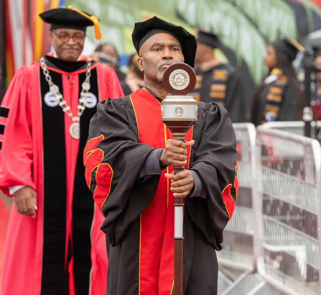 Professor in red and black regalia leads academic procession at graduation.
