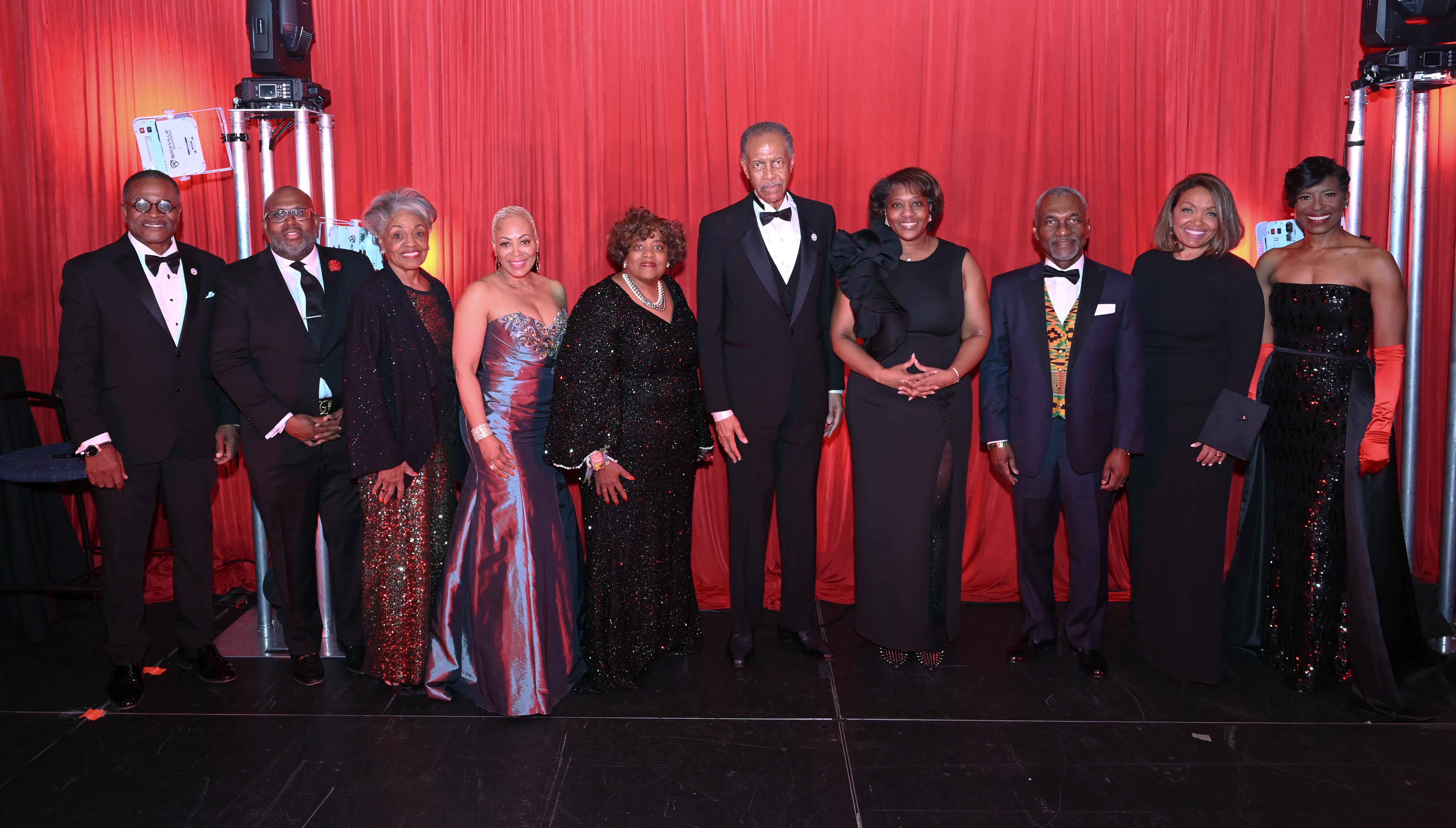Elegant group in formal attire against a red curtain backdrop.
