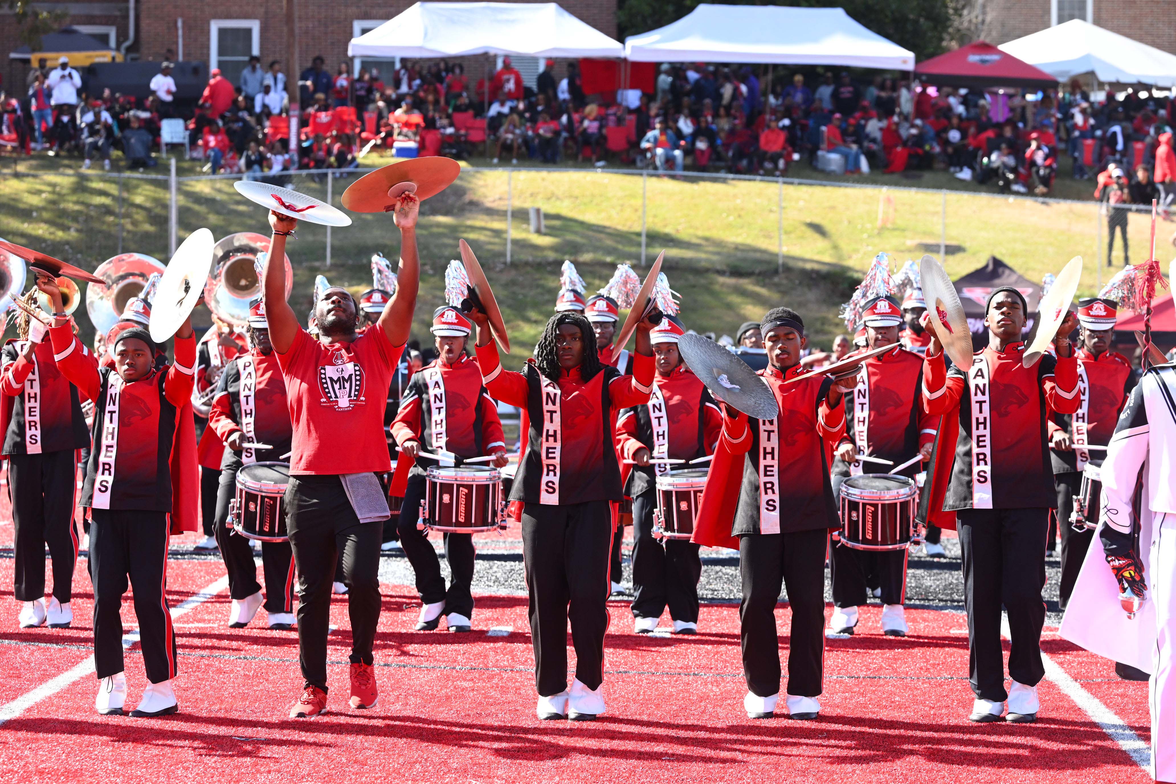 Marching band performing energetically on red track, with musicians in red uniforms.