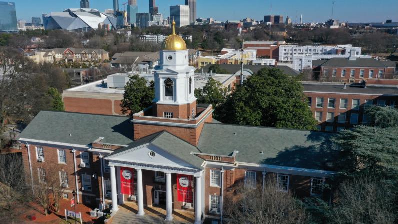 Brick building with a white bell tower, gold dome, and city skyline in the background.