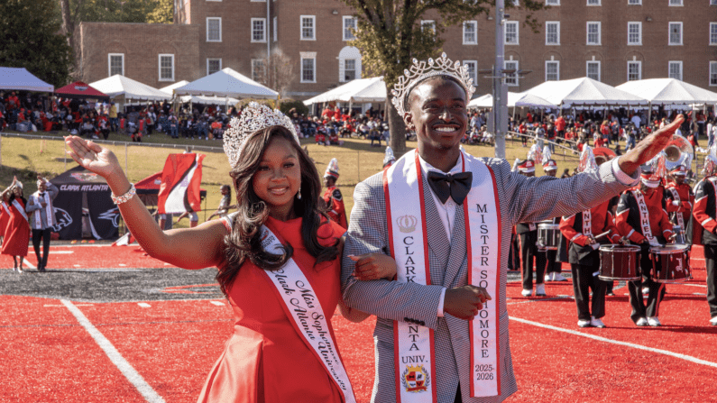 Couple wearing crowns and sashes on a red sports field, smiling, with crowd and band behind.