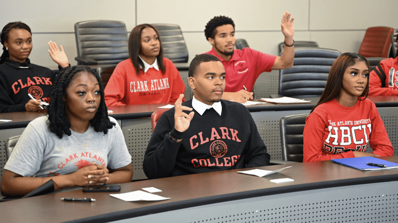 Students in a classroom, some raising hands, wearing collegiate sweatshirts.