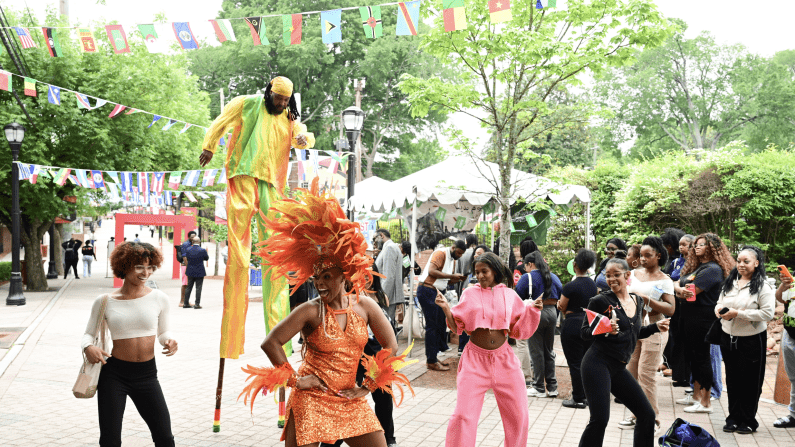Dancers in colorful costumes at an outdoor festival, with a stilt walker and flag decorations.
