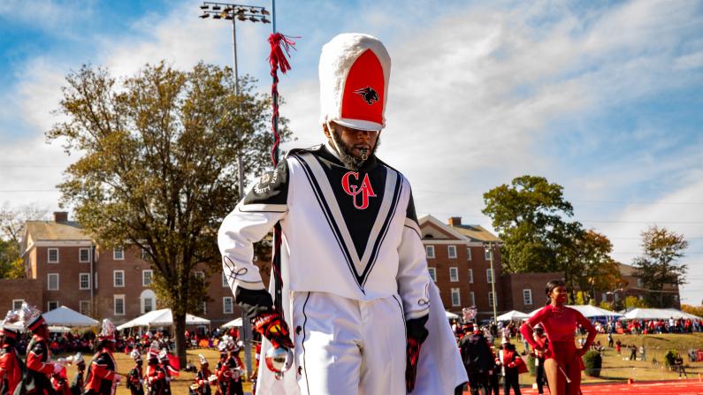 Marching band leader in tall white hat on red field, with trees and buildings behind.
