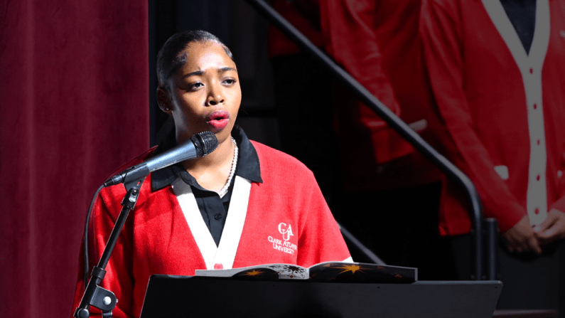 Woman in red sweater speaks at podium, choir in background.