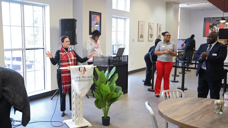 A person speaks at a podium; others listen in a modern room.