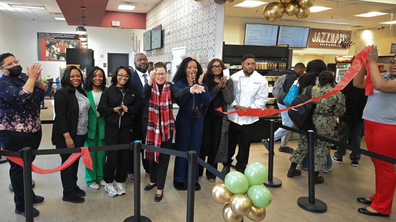 People gathered, cutting a red ribbon in a bakery shop opening event.