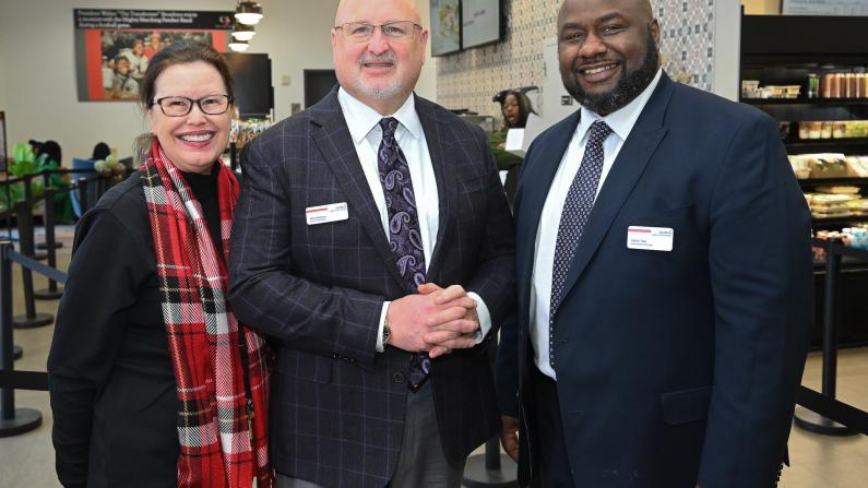 Three smiling people in business attire stand in a cafeteria setting.