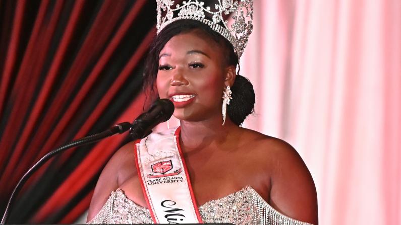 Pageant winner wearing a crown and sash speaks at a podium, smiling.