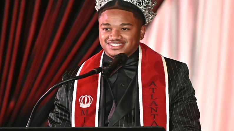 Man in a crown and red stole speaks at a podium.