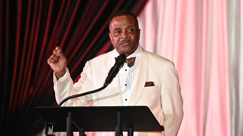 Man in a white suit speaking at a podium with a microphone.