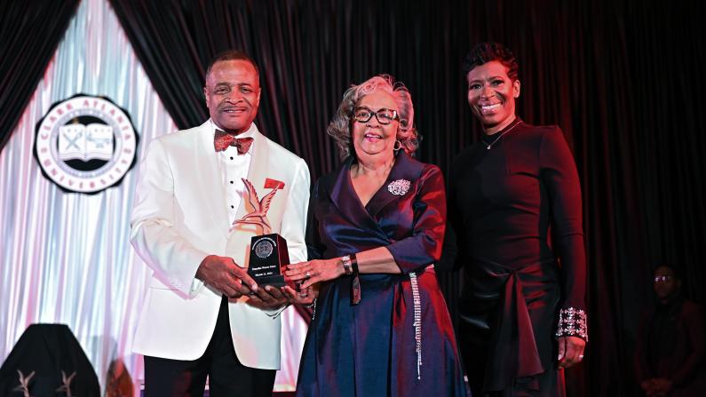 Three people in evening attire smiling, one holding an award.