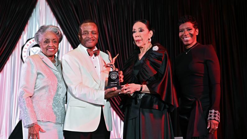 Four elegantly dressed people at an award ceremony, smiling on stage.