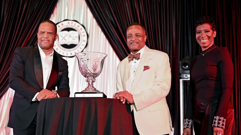 Three people in formal attire stand by a table with a trophy.