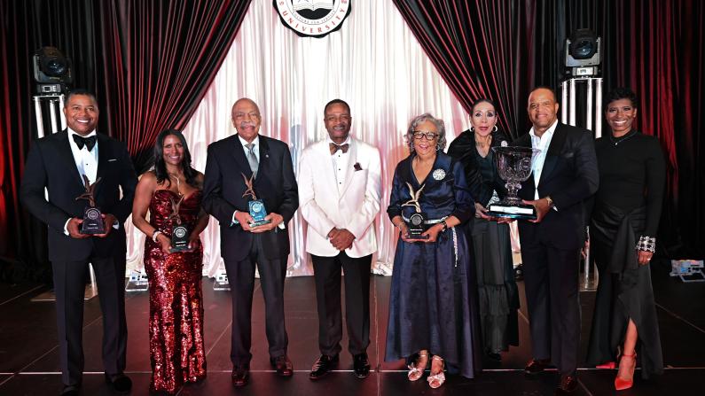 A group of people in formal attire holding awards on stage with curtains in the background.