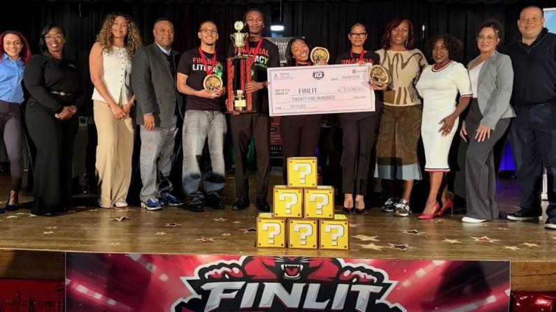 A group of people on stage holding a large check and trophies, with a colorful banner below.