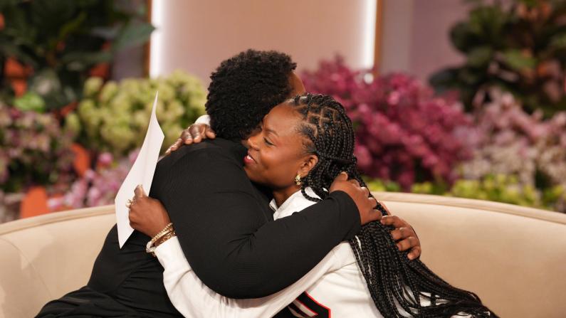 Two people embrace warmly on a talk show set with flowers in the background.