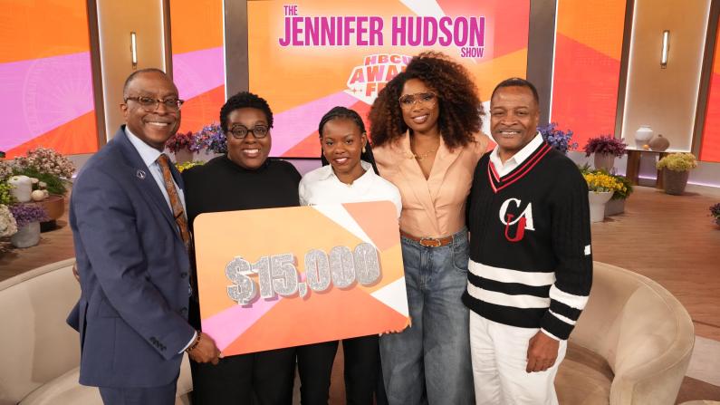 Five people smiling on a talk show set, holding a $15,000 sign.