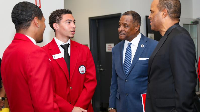 Three men in red blazers speak with two men in suits indoors.