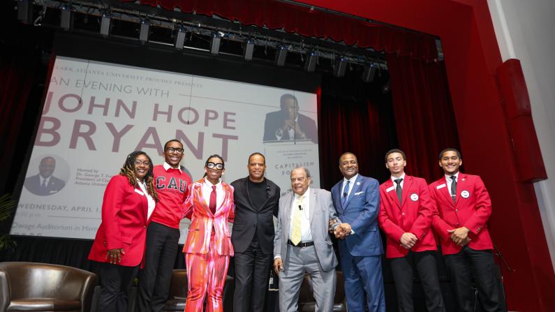 Group of people smiling on stage, red curtains, large screen backdrop.