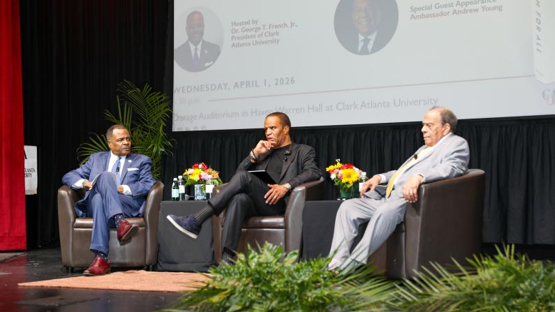 Three men seated on a stage panel, discussing, with a backdrop screen and plants around.