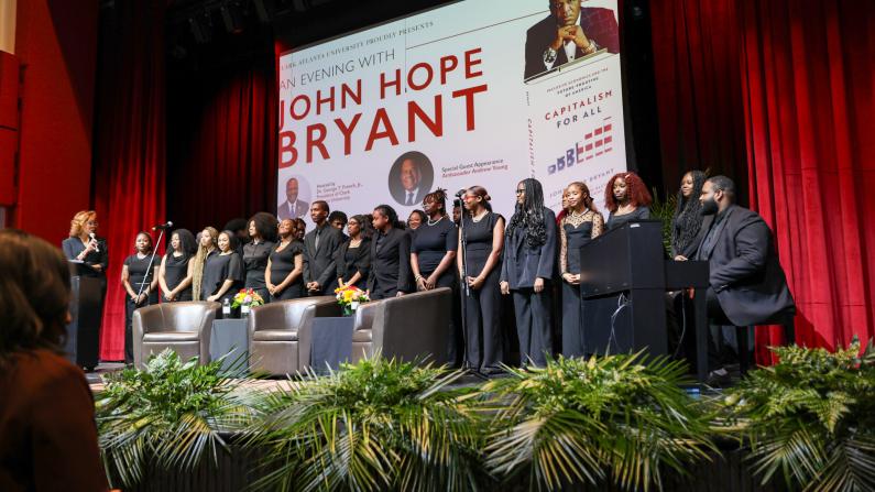 Audience facing a stage with people in black attire, red curtains, and greenery.