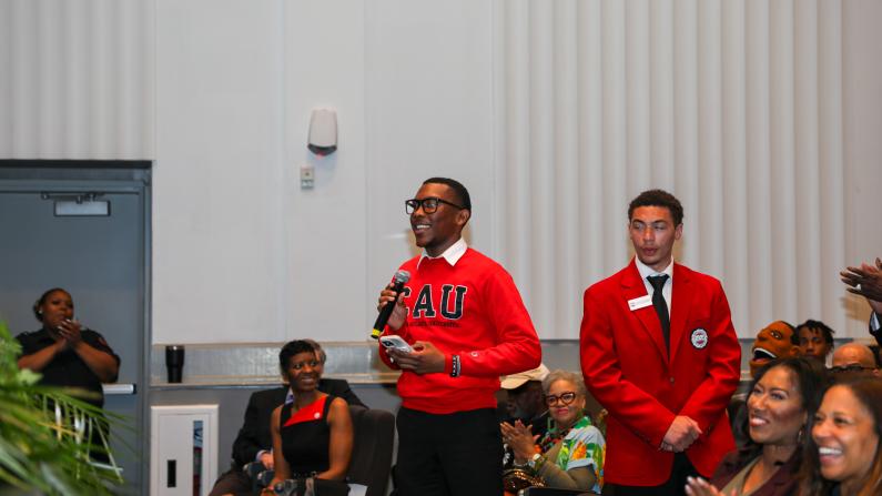 Man standing, speaking to an audience in a hall, wearing a red sweater with letters.