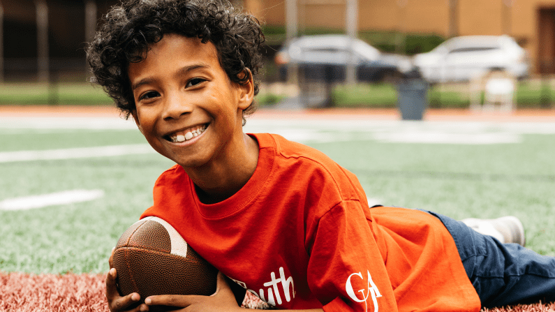 Smiling child in red shirt holds football on a field.