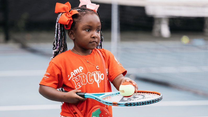 Young girl on tennis court holding racket and ball, wearing an orange shirt and bows.