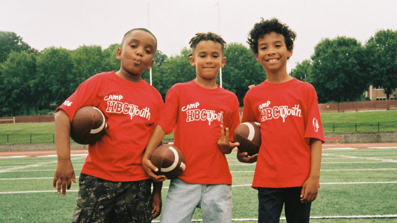 Three kids in red shirts holding footballs stand on a grassy field.