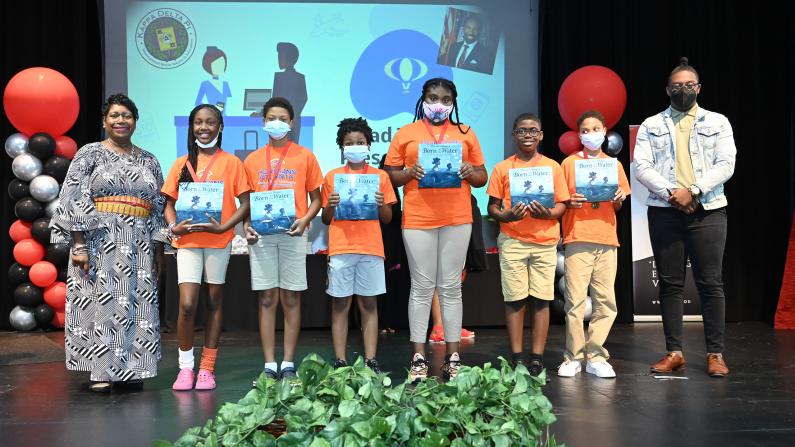 Children holding certificates on stage with two adults, balloons, and a screen in the background.