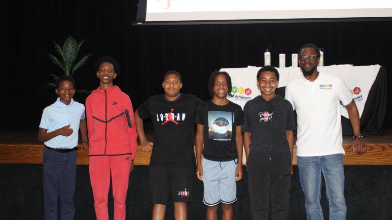 Group on stage, five smiling kids, one adult, casual clothing, indoor setting.
