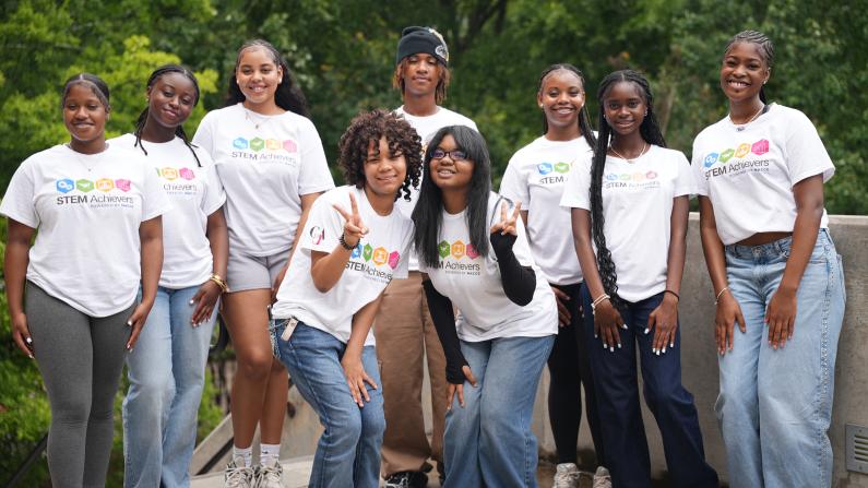 A group of diverse, smiling people in matching white T-shirts, posing outdoors.
