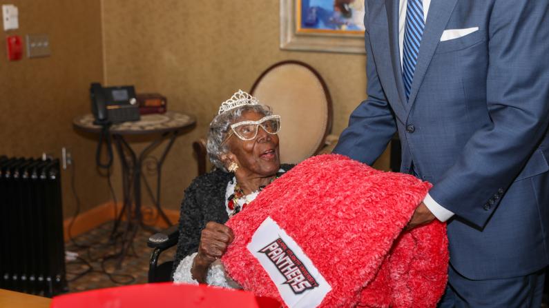 Elderly woman in a tiara receives red blanket from a man in a suit.
