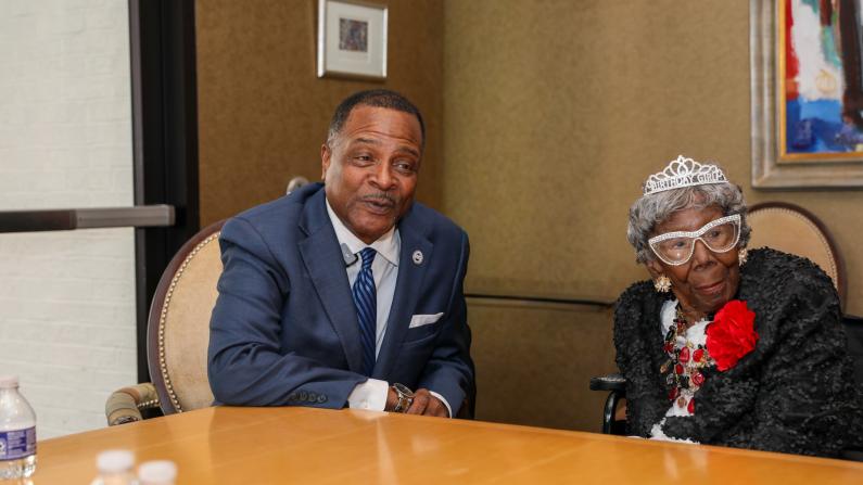 Man in blue suit sits next to an elderly woman with a crown and a red flower.