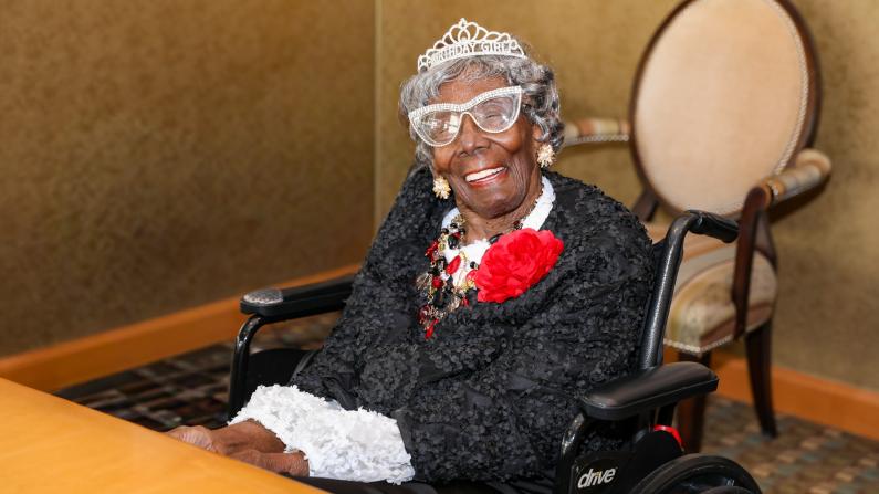 Elderly woman in a wheelchair wearing a tiara and red corsage, smiling warmly.