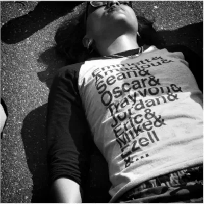 Person lying on pavement wearing a name-covered shirt, eyes closed. Black and white photo.