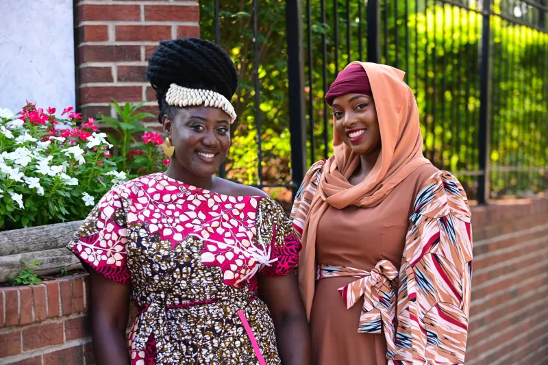 Two women smiling, dressed in colorful outfits, standing outdoors by a brick wall.