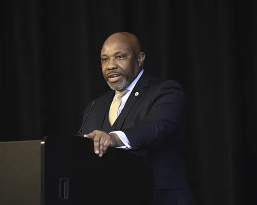 A man in a suit speaks at a podium against a dark background.