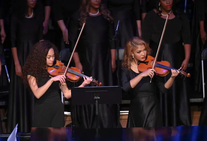 Two musicians in black dresses play violins on stage.