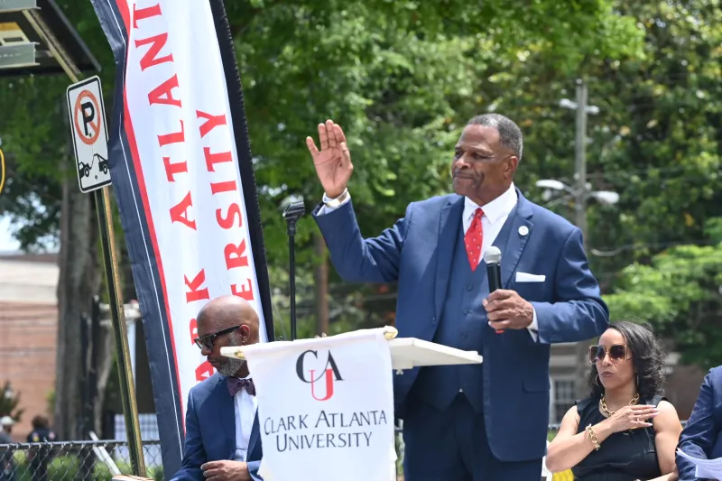 Man speaking at podium outdoors, wearing a blue suit and red tie.